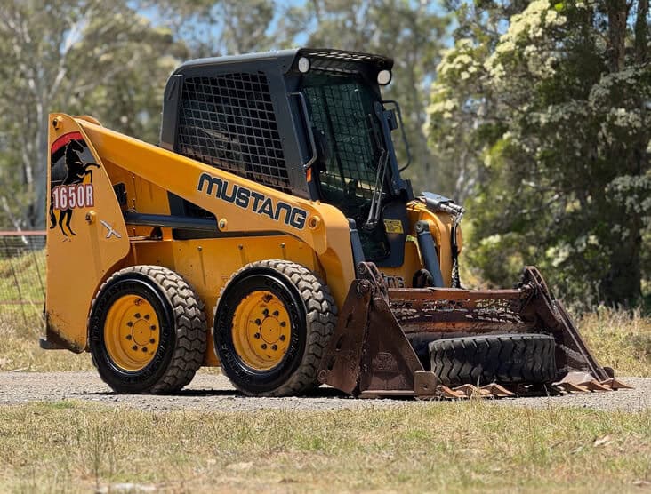 Yellow Mustang skid steer loader with attachment in outdoor setting.